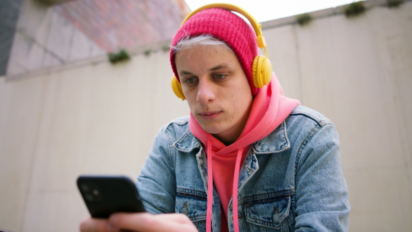 A low angle view of cool young man with headphones and smartphone outdoors in town, looking at camera.