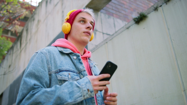 A low angle view of cool young man with headphones and smartphone walking outdoors in town.