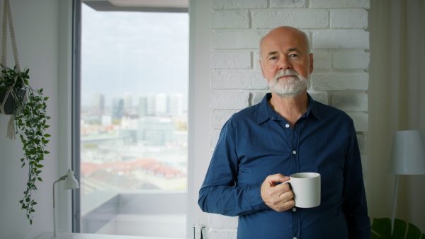 A senior man with coffee standing and looking at camera at home.