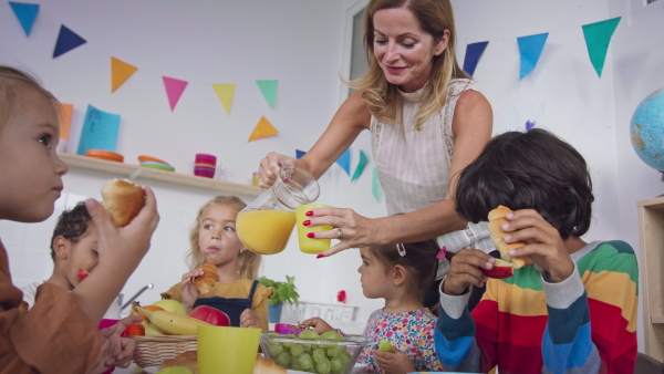 A group of little kids having snack indoors in nursery, teacher is assisting them.