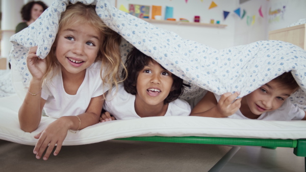 Preschool children in pajamas having fun hiding under a blanket indoors in nursery school, looking at camera.