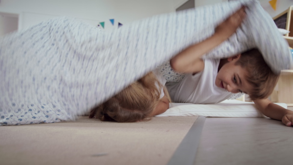 Preschool children in pajamas having fun hiding under a blanket indoors in nursery school.