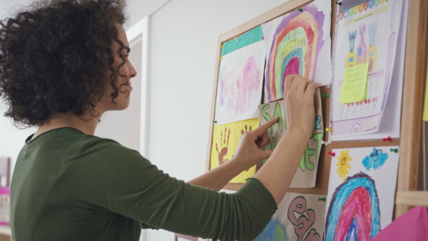 A teacher pinning posters and drawings on notice board at nursery school.