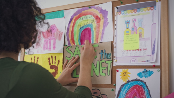 A teacher pinning posters and drawings on notice board at nursery school.