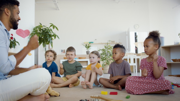 A group of small nursery school children with man teacher sitting on floor indoors in classroom, having lesson.