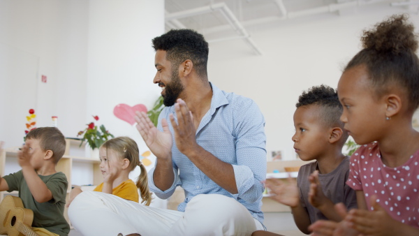 A group of small nursery school children with teacher sitting on floor, having music lesson and clapping.