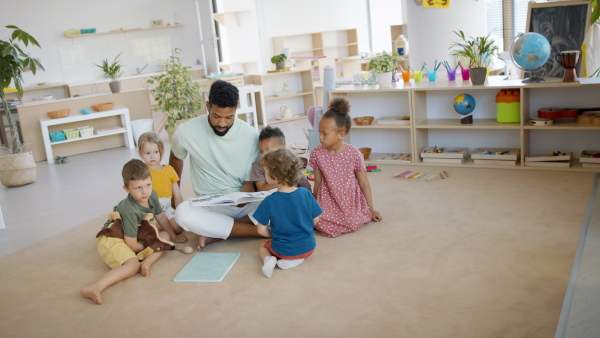 A group of small nursery school children with man teacher on floor indoors in classroom, reading book.