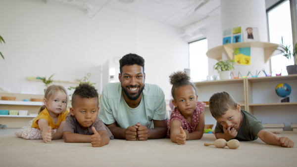 A group of small nursery school children with teacher on floor in classroom, looking at camera.