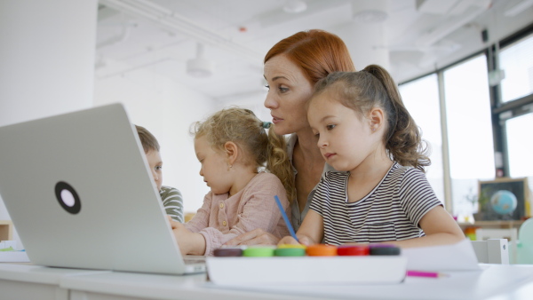 A group of small nursery school children with teacher indoors in classroom, using laptop.