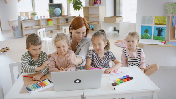 A group of small nursery school children with teacher indoors in classroom, using laptop.