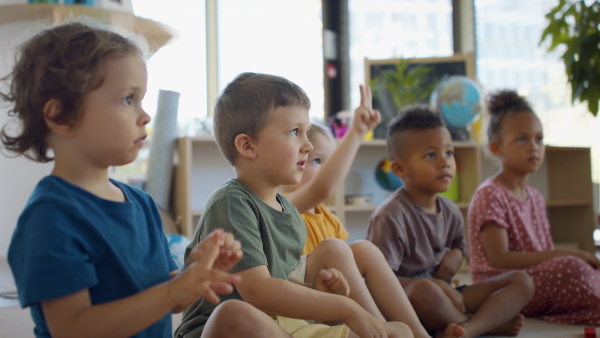 A group of small nursery school children sitting on floor indoors in classroom, raising hands.