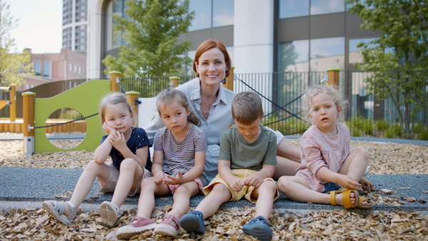 Group of small nursery school children with teacher sitting on playground, looking at camera.