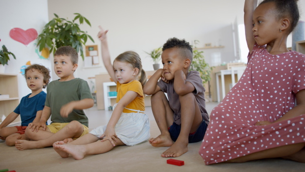 A group of small nursery school children sitting on floor indoors in classroom, raising hands.