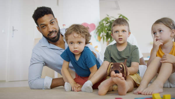 A group of small nursery school children with man teacher on floor indoors in classroom, looking at camera.