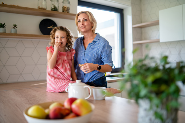 A happy cute small girl with mother indoors in kitchen at home.