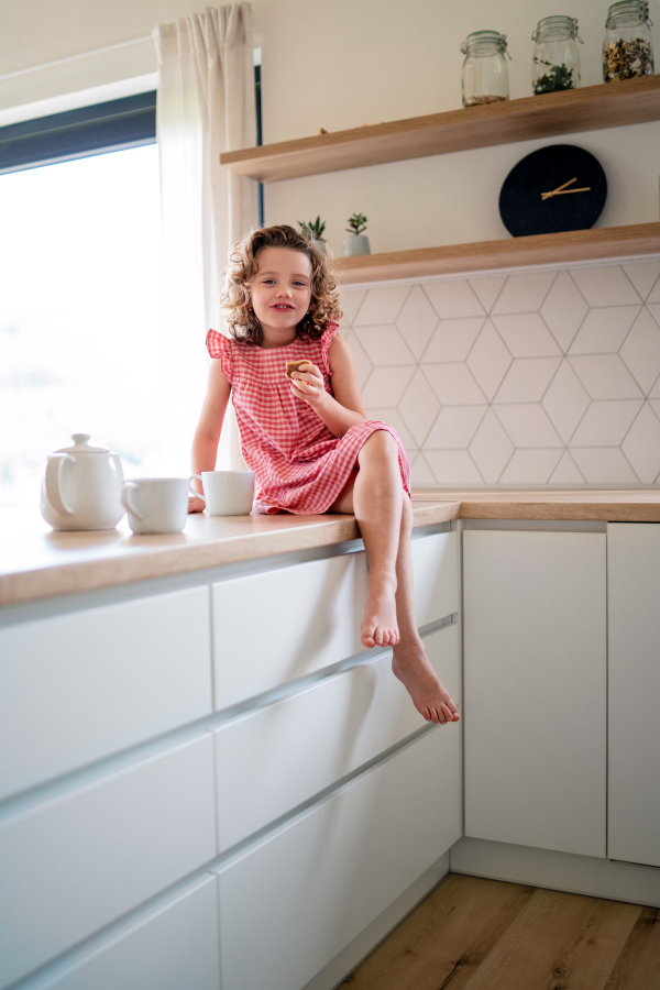 A beautiful cute small girl sitting on kitchen counter indoors at home, eating biscuits.