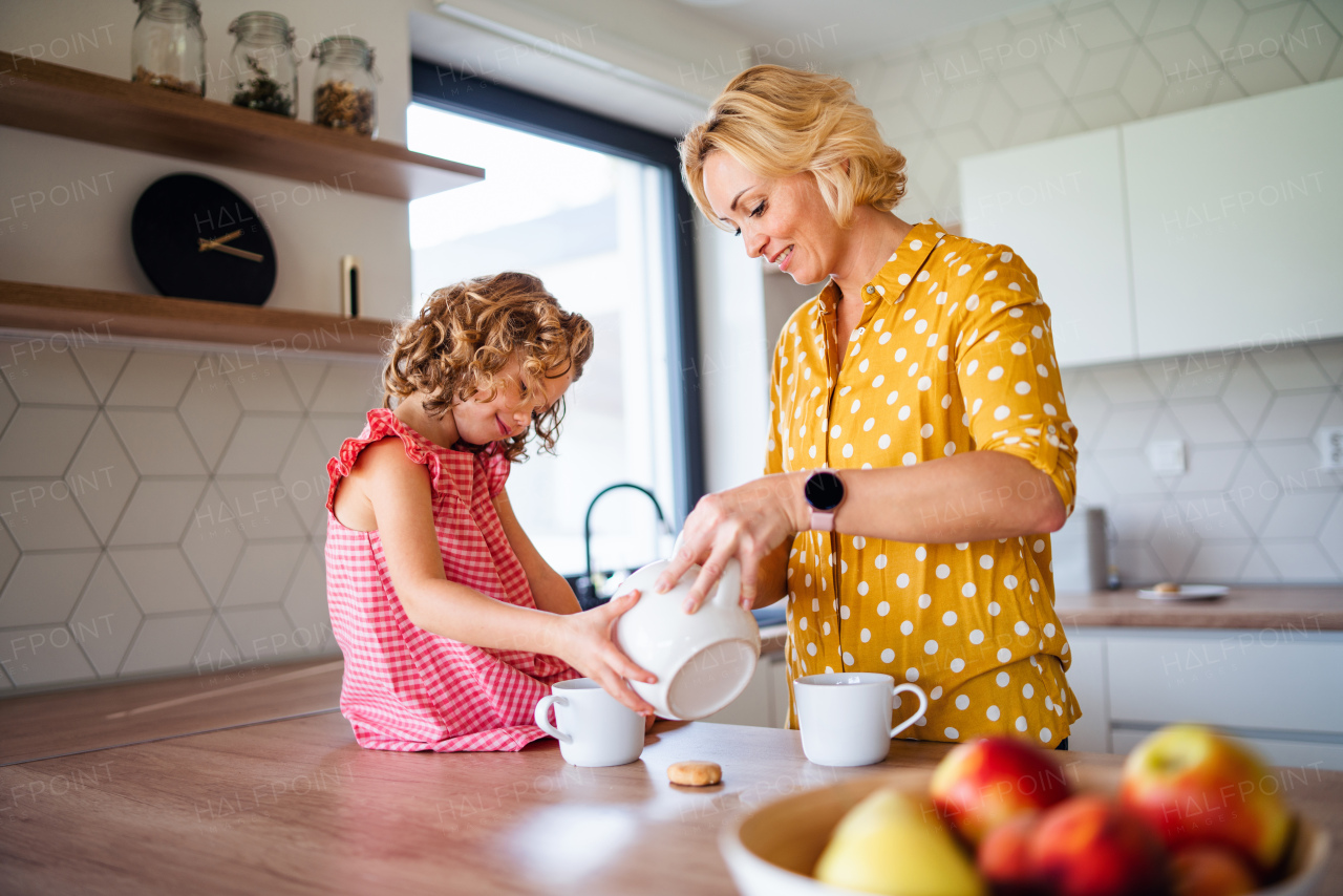 A happy cute small girl with mother indoors in kitchen at home, drinking tea.