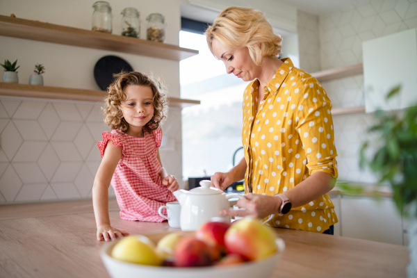 A happy cute small girl with mother indoors in kitchen at home, making tea.