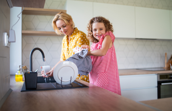 A cute small girl with mother indoors in kitchen at home, washing up the dishes.