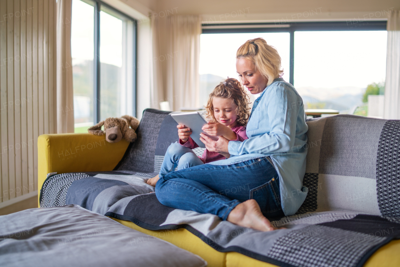 A front view of cute small girl with mother on sofa indoors at home, using tablet.