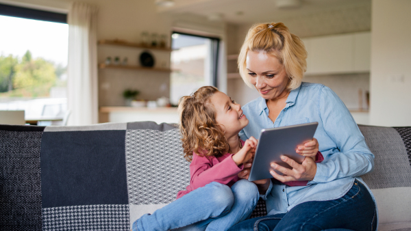 A front view of cute small girl with mother on sofa indoors at home, using tablet.