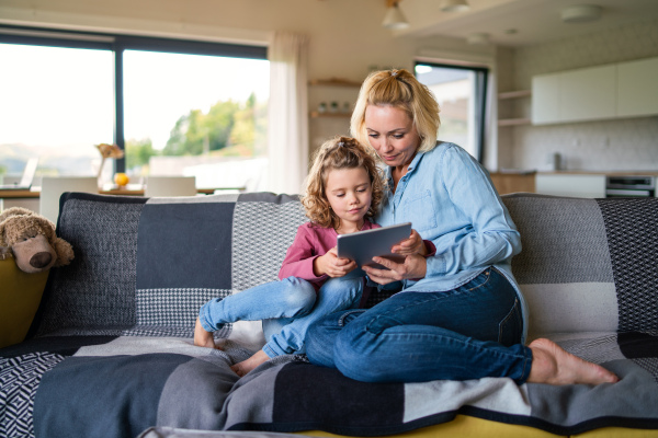 A front view of cute small girl with mother on sofa indoors at home, using tablet.