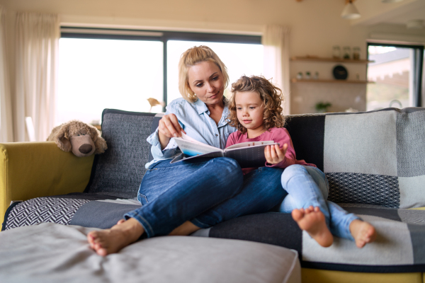 A cute small girl with mother sitting on sofa indoors at home, reading story book.