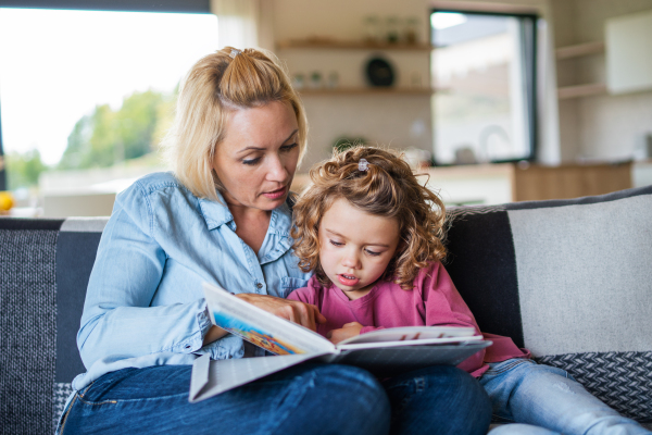 A cute small girl with mother sitting on sofa indoors at home, reading story book.