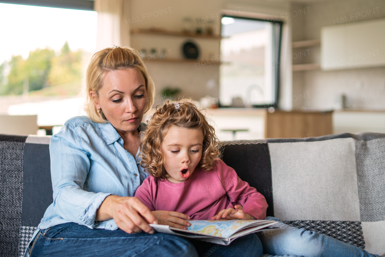 A cute small girl with mother sitting on sofa indoors at home, reading story book.