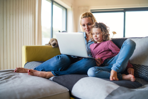 A cute small girl with mother sitting on sofa indoors at home, using laptop.