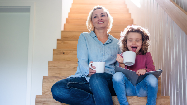 A front view of cute small girl with mother indoors at home, sitting and reading on staircase.