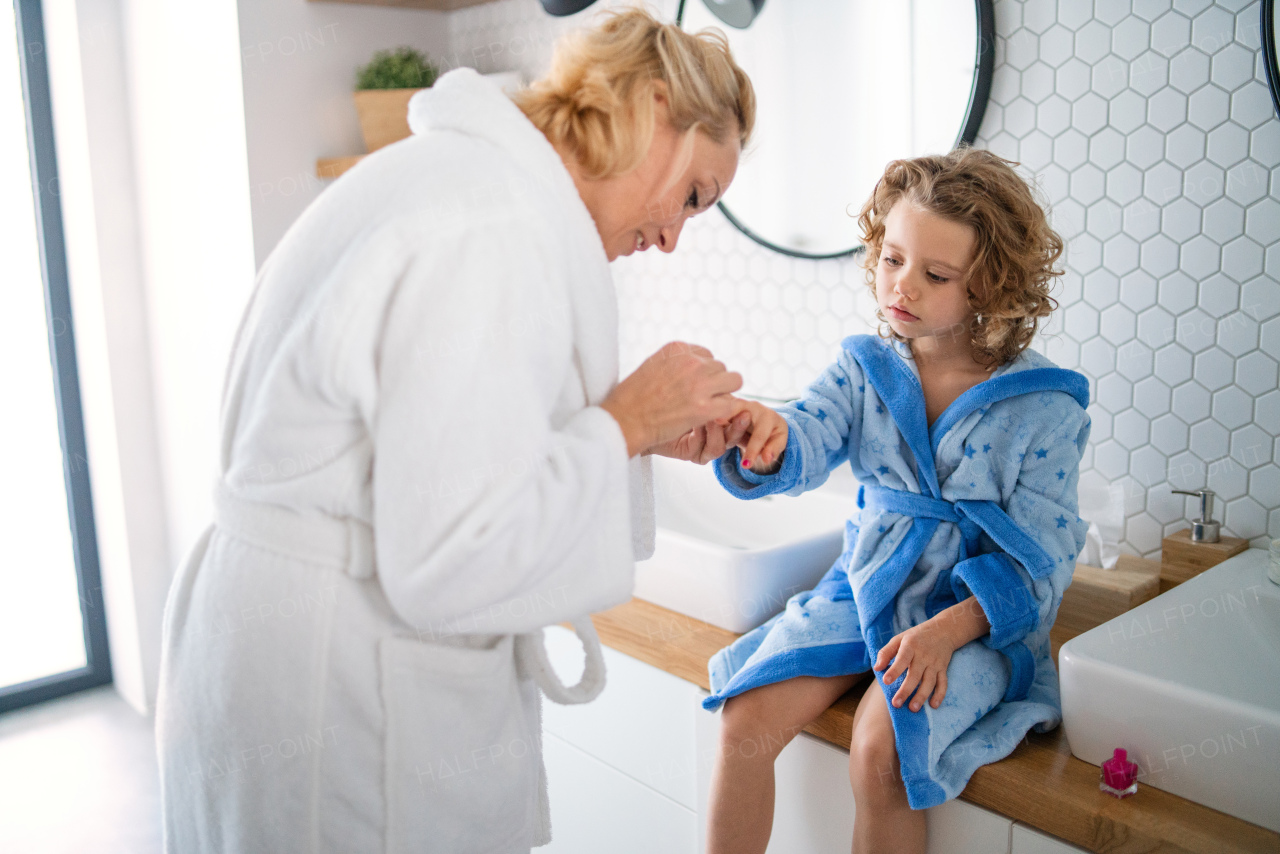 A happy cute small girl with mother in bathroom indoors at home, painting nails.