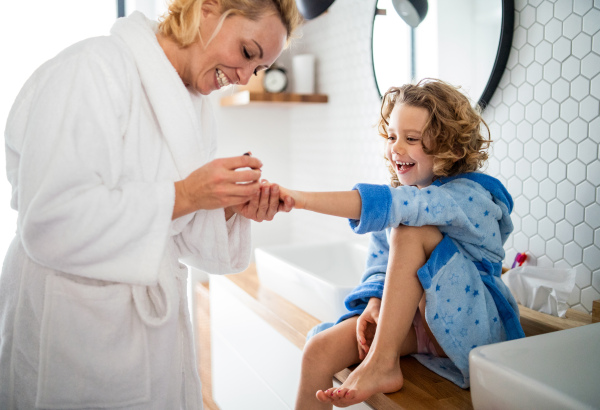 A happy cute small girl with mother in bathroom indoors at home, painting nails.