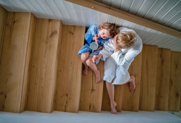 A top view of small girl with mother indoors at home, sitting on stairs in the morning.