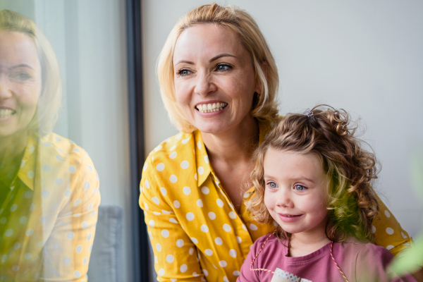 A cute small girl with mother indoors at home, looking out through window.