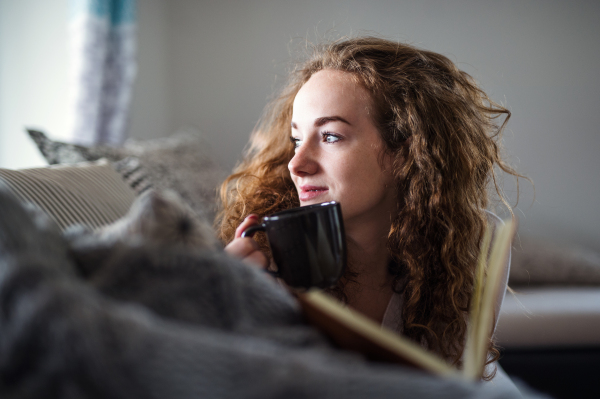 Young woman relaxing indoors at home with book and cup of coffee or tea.