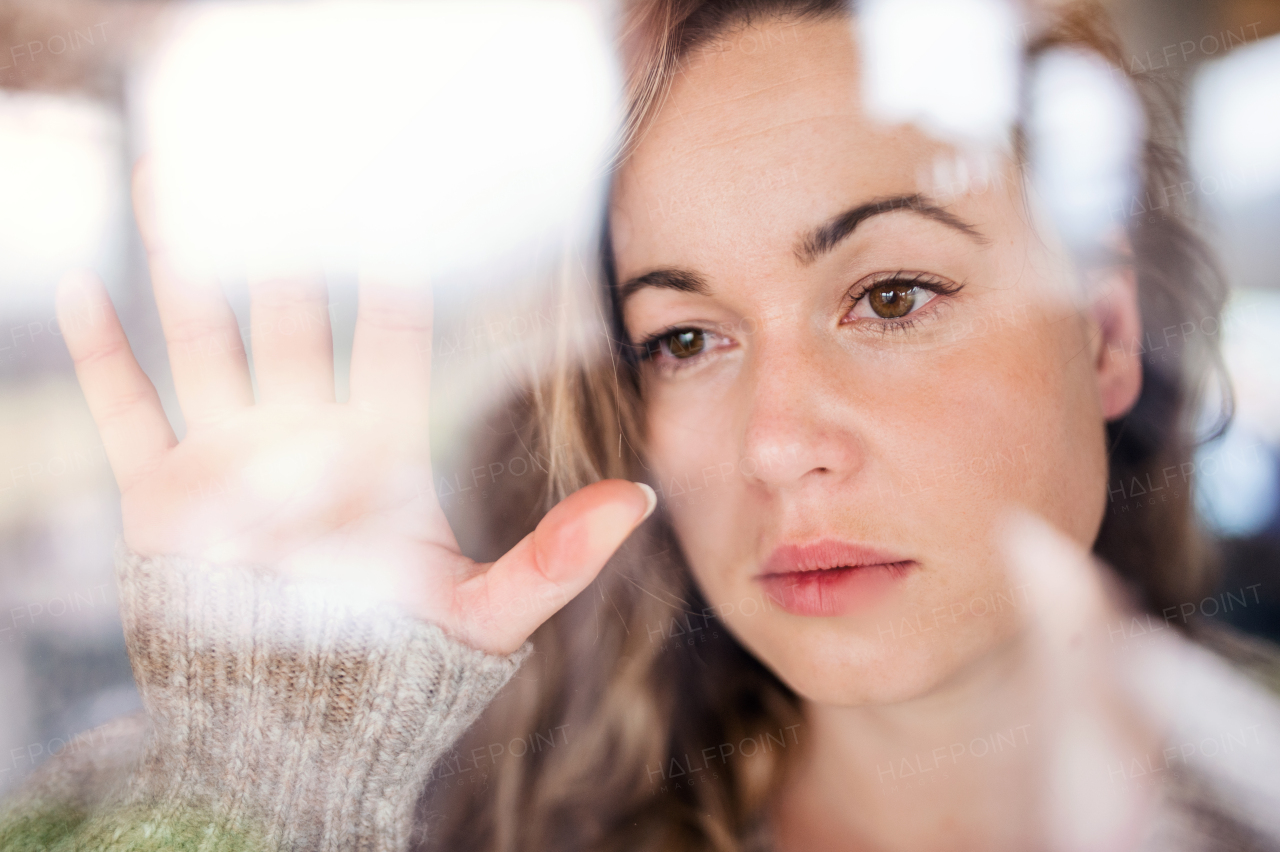 Young sad and depressed woman indoors by window at home. Shot through glass.