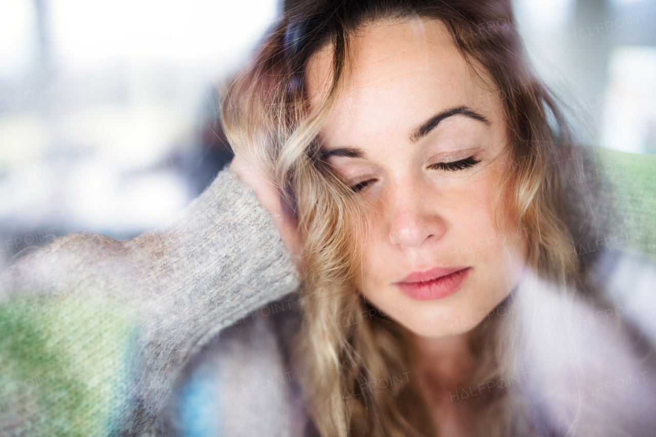 Young sad and depressed woman indoors by window at home. Shot through glass.