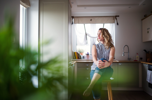 Front view of young woman sitting in kitchen indoors at home, relaxing.