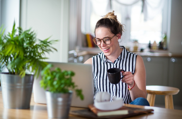 Portrait of young woman with laptop and coffee working indoors at home.