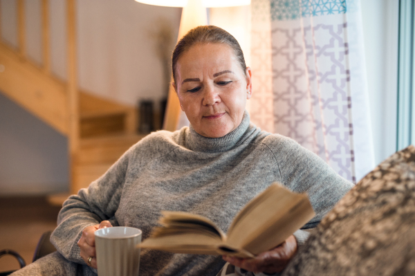 Front view portrait of senior woman sitting on sofa indoors at home, reading book.