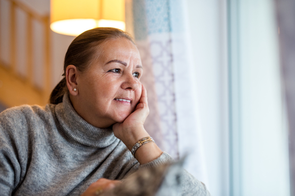 Close-up portrait of senior woman sitting by window indoors at home, looking out. Copy space.