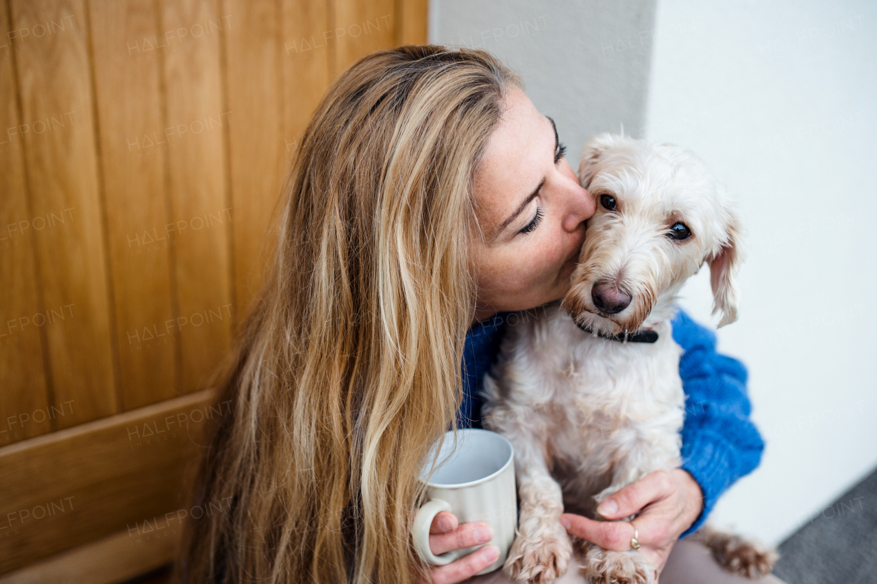 Mature woman relaxing outdoors by front door at home with coffee and pet dog, kissing.