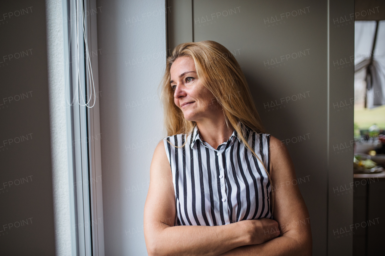 Front view portrait of woman standing by window indoors at home, resting.