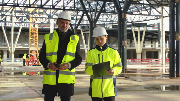 Man and woman engineers with walkie talkie walking on construction site, talking.