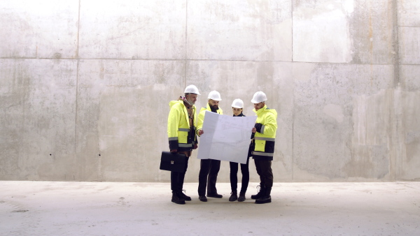 Group of engineers with blueprints standing against concrete wall on construction site.