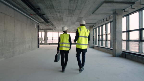 Rear view of man and woman engineers with bag walking on construction site, talking.