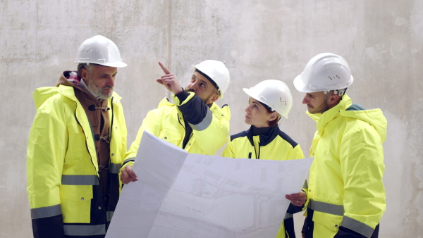 Group of engineers with blueprints standing against concrete wall on construction site.