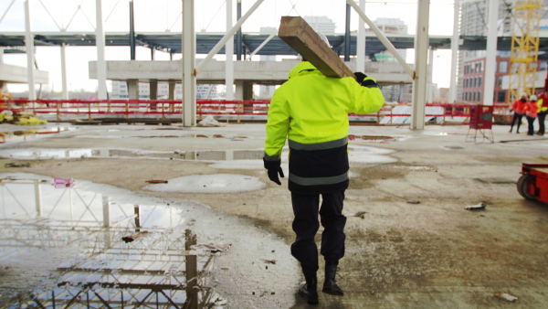 A rear view of man worker walking outdoors on construction site, carrying beam.