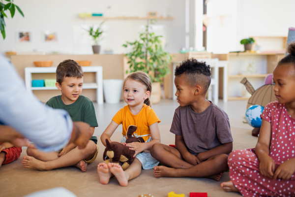 A group of small nursery school children sitting on floor indoors in classroom, listening to teacher.